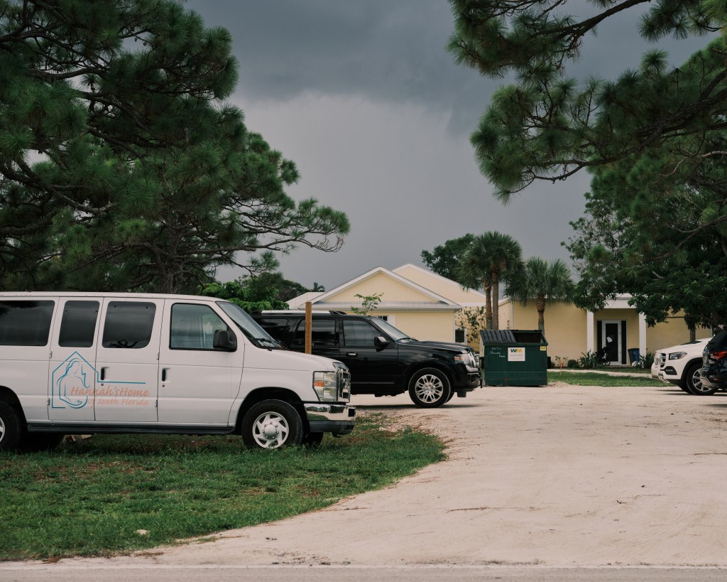 Cars are parked along a long driveway leading to a yellow single-story home. One van bears a logo for Hannah's Home of South Florida.
