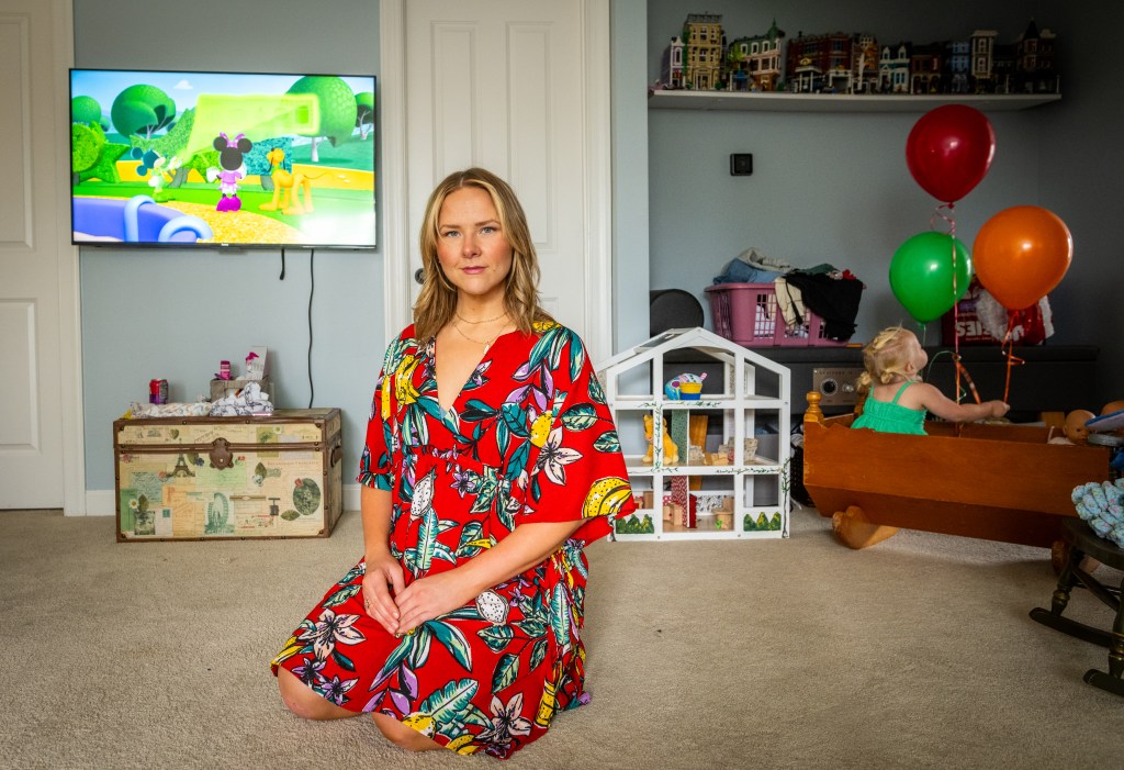 Rachel Hunt sits on the floor of a room filled with children's toys, with a Disney cartoon showing on a TV behind her. Her daughter sits in a wooden crib nearby.