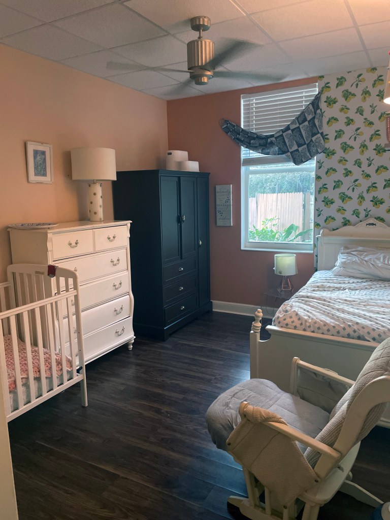 A pink-walled room holds a single bed, a crib, a pair of dressers, and a padded rocking chair.