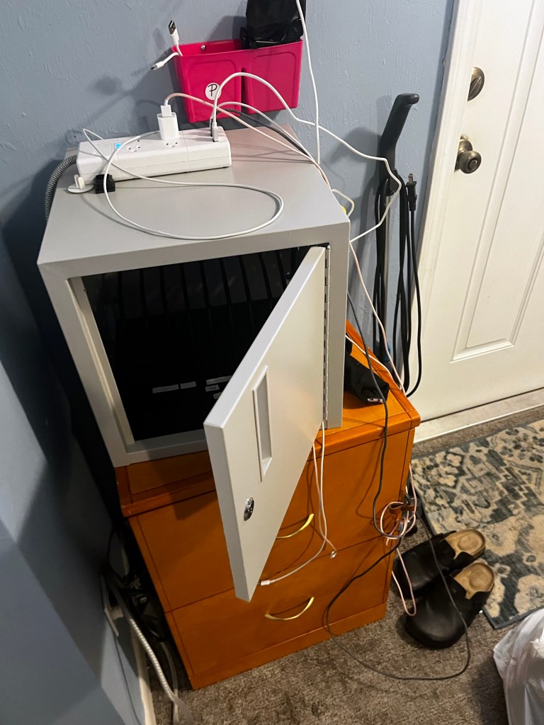 A small safe with a key lock sits on a nightstand. Charging cables dangle from a surge protector sitting on top.