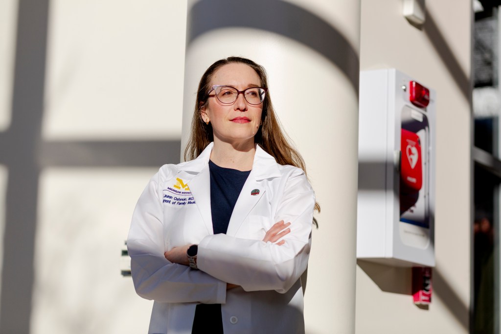 A White female doctor poses for a portrait at a hospital. She is wearing glasses, a black blouse, and a white lab coat.